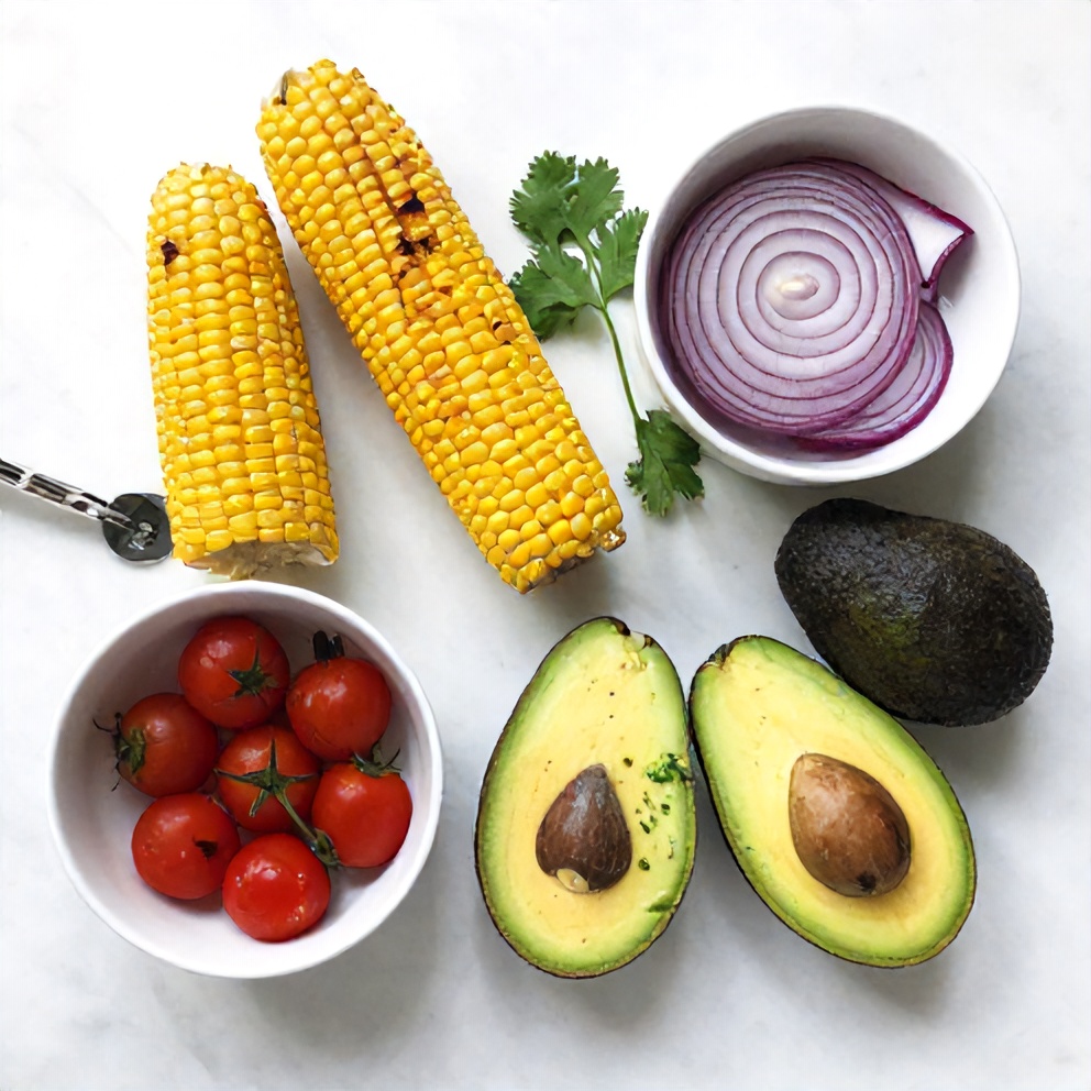 Raw ingredients for Fresh Grilled Corn Avocado Salad on a white marble surface